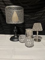 Five candle holders displayed on black fabric background. Includes black metal holder with mosaic shade, two clear glass votive holders with circular cut design, silver mercury glass holder, and small silver metal holder with crystal bead decoration.