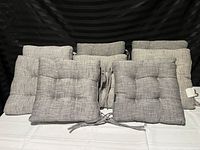 Eight gray tufted fabric chair cushions with ties displayed on a white surface and black backdrop.