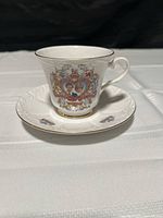 Tea cup and saucer set showing the detailed front design featuring the portraits of Charles and Diana in a heart-shaped frame with heraldic and floral designs and gold rim details.