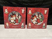 Two boxed sets of four St Nicholas Collection Christmas-themed dinner plates showing illustrations of Santa and Christmas trees on red boxes