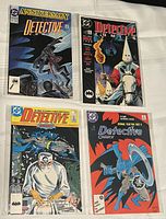 Four Detective Comics comic books in plastic sleeves laid on white fabric. Covers show Batman-related themes and characters in colorful and graphic art style.