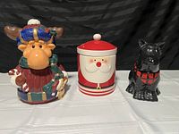 Photo of three ceramic Christmas-themed cookie jars: moose with blue crown and candy canes, round Santa Claus with red lid, and black dog figure with plaid sweater.
