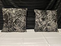 Photo showing two square pillows with black and white swirling abstract pattern on fabric, placed on white surface with dark backdrop