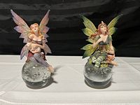 Both fairy figurines shown side by side from the front, depicting details of the wings, outfits, and glass ball bases.
