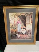Photo of framed art print showing a woman standing under an arch with a child and cat in a warm outdoor setting.