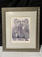 Framed art print showing three classical urns in monochrome inside an ornate metallic silver-gray embossed frame with cream matte.