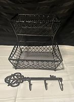 Black metal two-tiered basket positioned above black cast iron hanging key with hooks on white surface against black background
