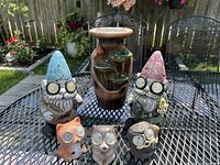 Photo showing six ceramic solar garden figurines: two gnomes with hats and glasses, three owls with glasses, plus a decorative ceramic vase with leaf design.