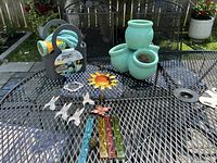 Photo showing various garden items on black metal mesh table, including garden hose on holder, turquoise ceramic pots, metal garden stakes shaped like animals and fairies, solar flower light.