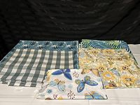 Four assorted tablecloths laid out flat showing different patterns and colors including butterflies, checks, leaves, and flowers.
