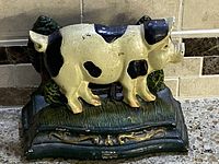 Front angle view of cream pig with black spots cast iron holder on green base with brown fence backdrop
