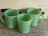 Set of three jadeite green glass mugs arranged side by side on a countertop, showing shape and color.