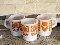 Three white milk glass mugs with orange floral and geometric designs presented on a counter in front of beige brick backsplash.