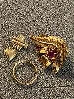 Three jewelry pieces on grey surface: 18K gold clip-on earring with red stones, 10K gold small hoop, 10K gold cross stud with heart charm