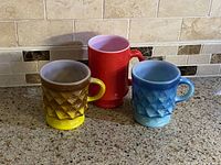 Three vintage milk glass mugs: yellow/brown diamond pattern, blue diamond pattern, red smooth mug with pink interior, shown on countertop