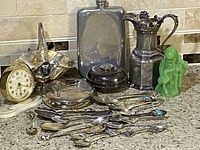 Lot showing vintage clock, silverplate pitcher, flask, lidded dish, green figurine, and assorted spoons spread on countertop.