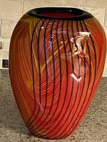 Front angle of oval Murano style vase showing red, yellow, black and blue swirl stripes and dark rim.