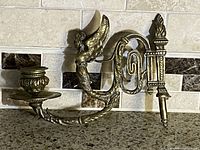 Full view of the brass wall-mounted candle holder sconce against tiled wall, showing candle cup, arm, winged figure, and torch detail.