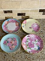 Four Royal Albert Miranda Kerr Gratitude appetizer plates shown from top view arranged in square formation, showcasing floral and butterfly patterns with gold rims
