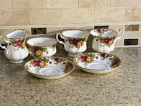 Five piece Royal Albert Old Country Roses bone china tea set displayed on granite countertop. Includes two cups, two saucers, cream pitcher, and sugar bowl. Gold trim and floral motifs visible.