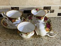 Full view of two Royal Albert Old Country Roses teacups resting on matching saucers, along with creamer and sugar bowl showing gold trim and detailed multicolor floral rose pattern.