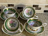 Photo showing a set of 4 Royal Stafford bone china teacups with matching saucers arranged on a countertop, highlighting green and gold rims with floral interior designs.
