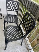 Two black metal lattice outdoor chairs side by side on balcony with a view of railing and greenery below.