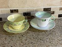 Photo shows the two tea cup and saucer sets side by side on a countertop, highlighting the shapes and colors of each cup and saucer.