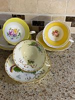 Three vintage bone china teacups and saucers arranged on a countertop showing floral and bird patterns with yellow trim.