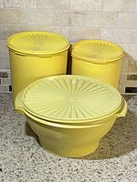 Photo showing three yellow plastic Tupperware containers with matching lids featuring a radiating sunburst pattern, placed on kitchen counter against tile backsplash.