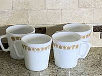 Four white milk glass mugs with a gold Butterfly Gold decorative band near the rim, placed on a tiled surface.