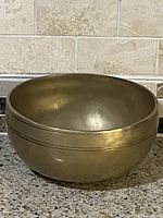 Photo showing the round brass singing bowl with a matte finish from a side angle, placed on a speckled countertop with a tiled backsplash.