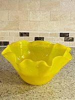 Front and side view of the vintage yellow glass fruit bowl placed on a granite countertop with tiled backsplash, showing the ruffled scalloped edge and translucent yellow glass.