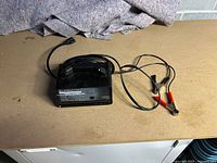 Photo showing Eliminator 12V intelligent battery charger with power cord and red and black alligator clips on a work surface.