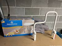 Photo of the assembled white metal bath safety bar with curved handle, clamps on each side, set against a concrete block wall with the AquaSense bath seat box in the background.