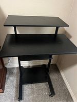 Full view of the black computer desk showing main desk area and raised secondary platform, highlighting the height adjustable design and caster wheels under base.