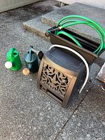 Photo showing hose reel, two watering cans, and planter on pavement with steps in background
