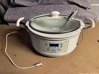 View of the full slow cooker showing the white oval body, side handles, transparent glass lid with a knob, and attached temperature probe cable.