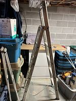 Photo showing tall, aluminum folding ladder with paint splatters and signs of use, partially opened and standing upright in a basement.