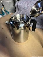 Photo of the percolator with lid open showing the interior filter basket and polished stainless steel exterior with black handle.