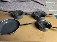 Photo showing the set of cookware including one frying pan and three covered pots, laid out on a cardboard surface with protective blankets in background.
