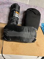 Three packed camping items: one sleeping bag and two pillows on a flat surface.