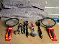 All seven kitchen items displayed on table against a cinderblock wall background, showing strainers, utensils, meat tenderizer, and packaging.