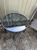 Round glass-top table with metal frame on gravel outdoor surface.