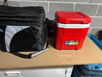 Two drink coolers on a table: a black soft-sided cooler bag and a red and white Igloo cooler