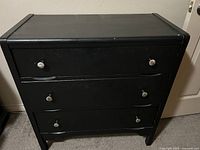 Front view of the black solid wood three-drawer dresser showing metal knob handles and visible wear.