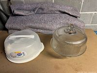 Three cake containers on a table with a folded blanket in the background. Two are plastic with translucent lids and white bases; one is a glass container with a ribbed dome lid and pedestal base.