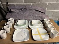 Two matching 16-piece Corelle dinnerware sets including plates and mugs, floral pattern and white with yellow ribbon design.