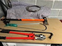 Axe with orange handle, crowbar, toilet snakes, and bolt cutters displayed on table.