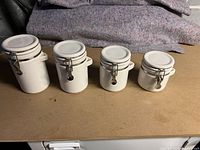Four white ceramic canisters of varying sizes arranged in descending order from left to right on a flat surface with a cloth backdrop.